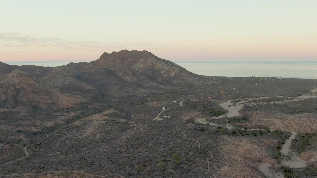 Wide aerial frame in Cabo Pulmo showing a central mountain mass, dry washes, dirt tracks, low scrub, distant sea, and no visible people.