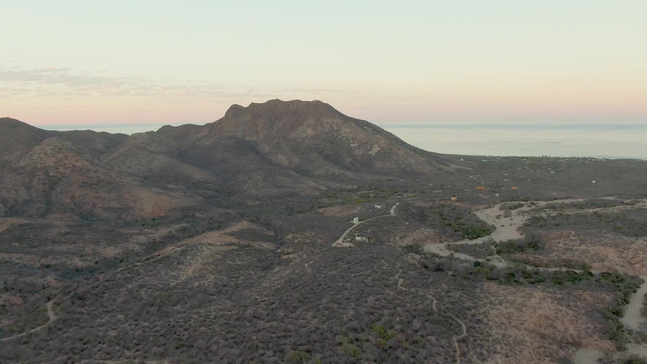 Aerial frame over Cabo Pulmo arid hills with a large mountain left of center, pale ocean horizon, winding roads, dry washes, and no visible people.
