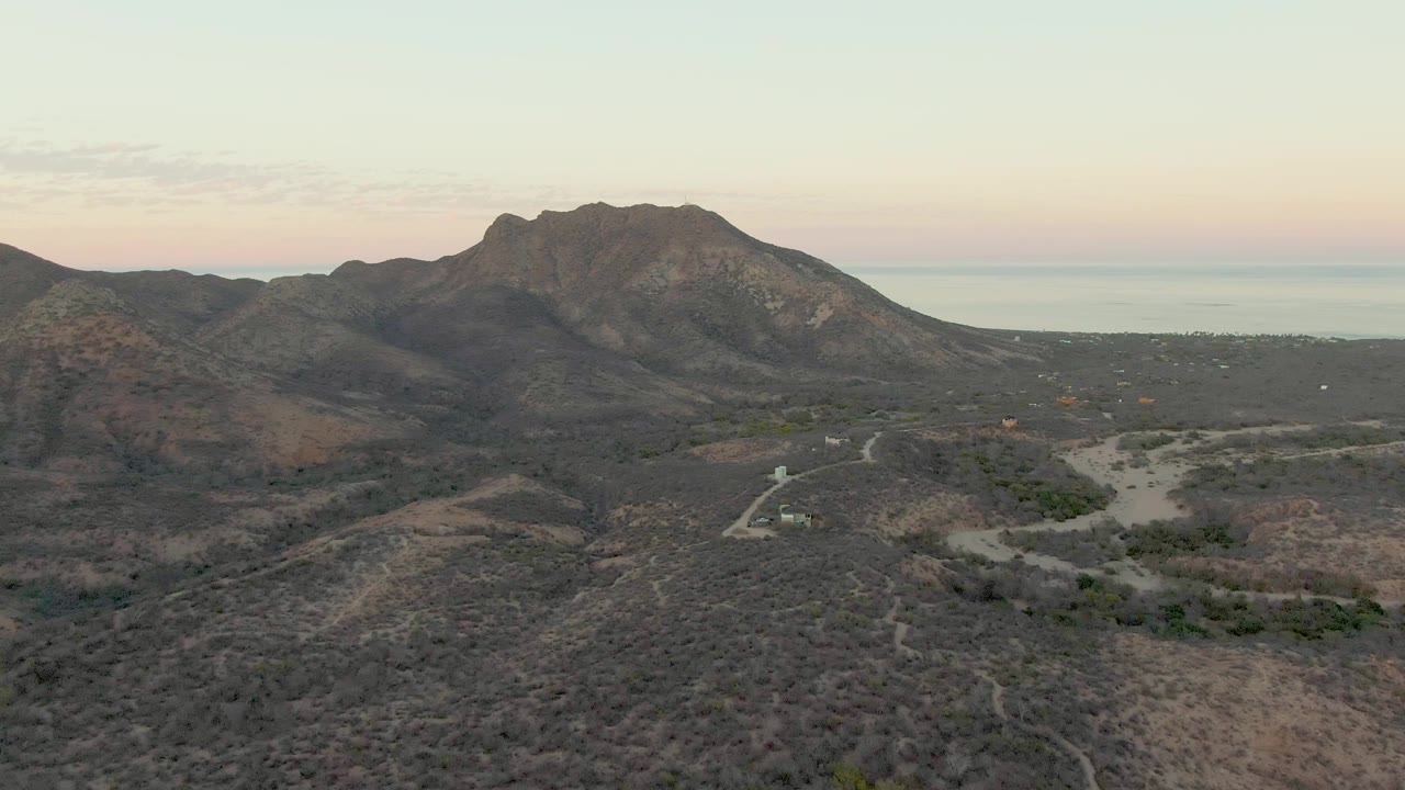 Aerial frame over Cabo Pulmo with the dirt road and small vehicle slightly farther along the valley, surrounded by scrub-covered hills, mountain ridges, distant sea, and no visible people.