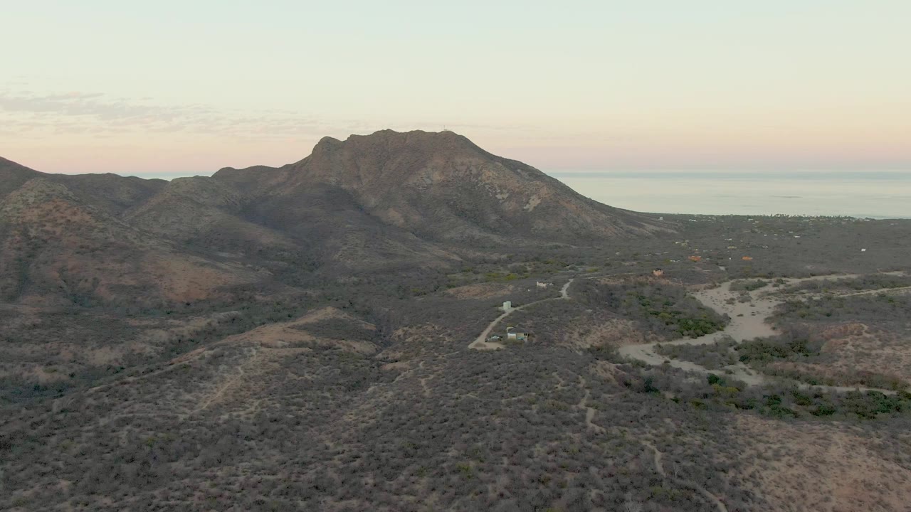 Mid-clip aerial view in Cabo Pulmo showing a small vehicle on a pale dirt road below dry rolling hills, dark mountains, a narrow sea horizon, and no visible people.