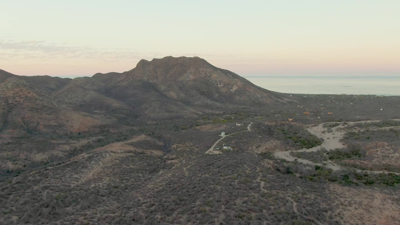 Later aerial frame over Cabo Pulmo with the mountain mass shifted across the background, scrubby hills in the foreground, the dirt road visible in the valley, and no visible people.
