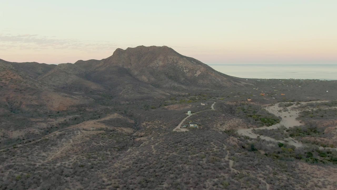 Dusk aerial view of a mountain ridge, dry hills, winding road, and distant ocean.
