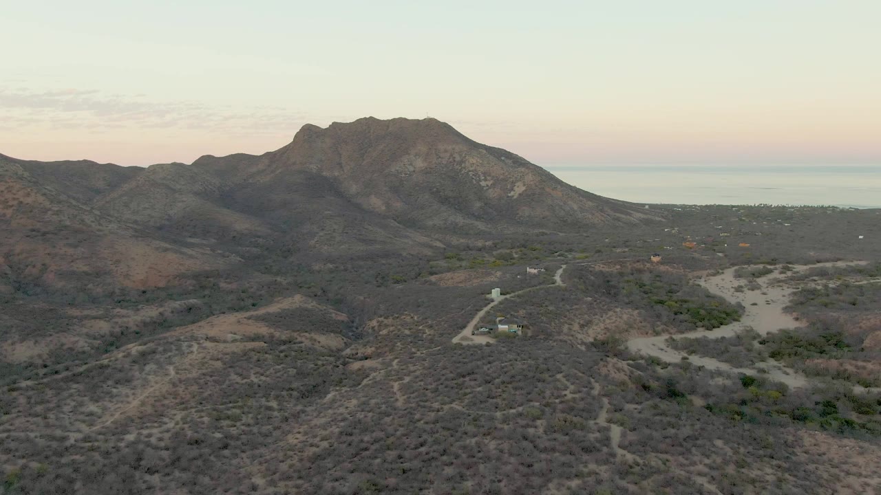 Second frame from the Cabo Pulmo aerial view, with the ridge slightly more centered over muted desert vegetation, the pale road in the lower terrain, and no visible people.