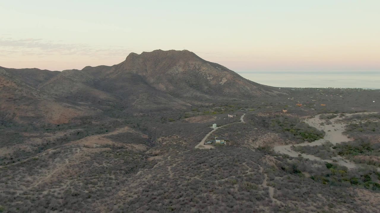Third frame looking across Cabo Pulmo scrub hills toward the dark ridge, with the road tracing through the lower left area, the ocean beyond, and no visible people.