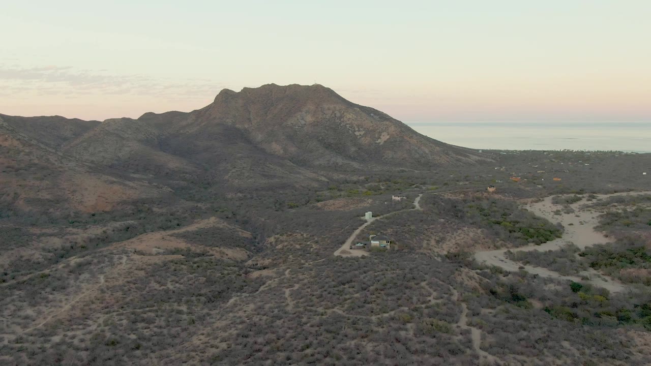 Fourth frame in the Cabo Pulmo clip, with a rugged mountain ridge spanning the middle, dry hillside texture in the foreground, the ocean horizon beyond, and no visible people.