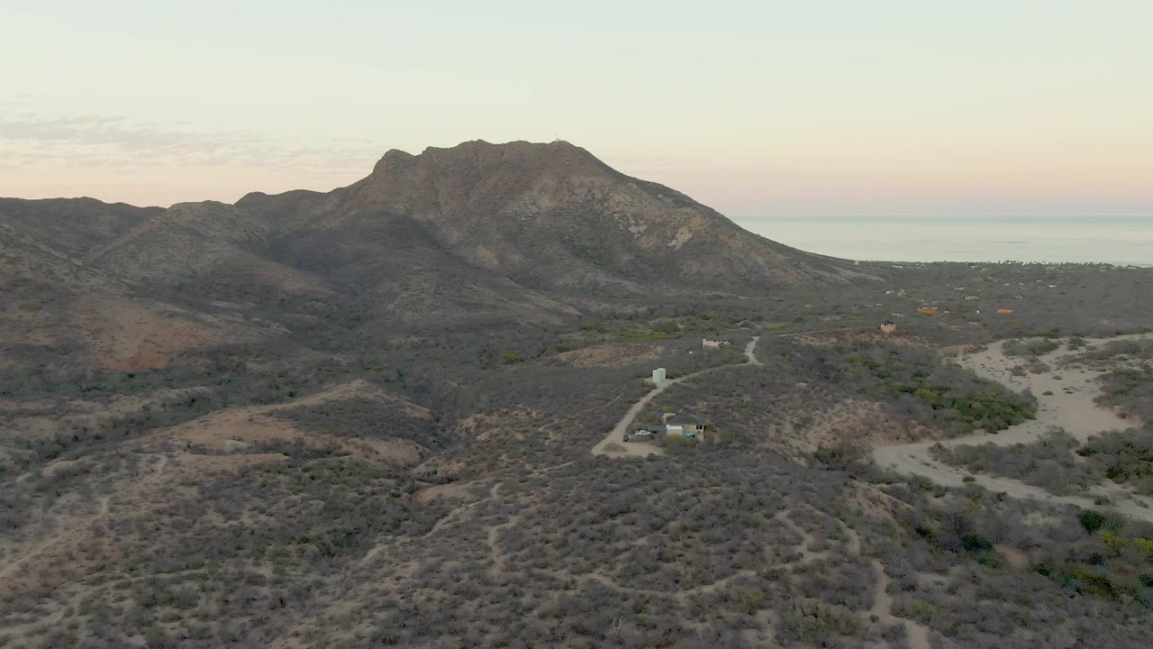 Wide aerial frame over Cabo Pulmo desert terrain, with a pale dirt road, sparse scrub, distant mountains, a faint sea horizon, and no visible people.