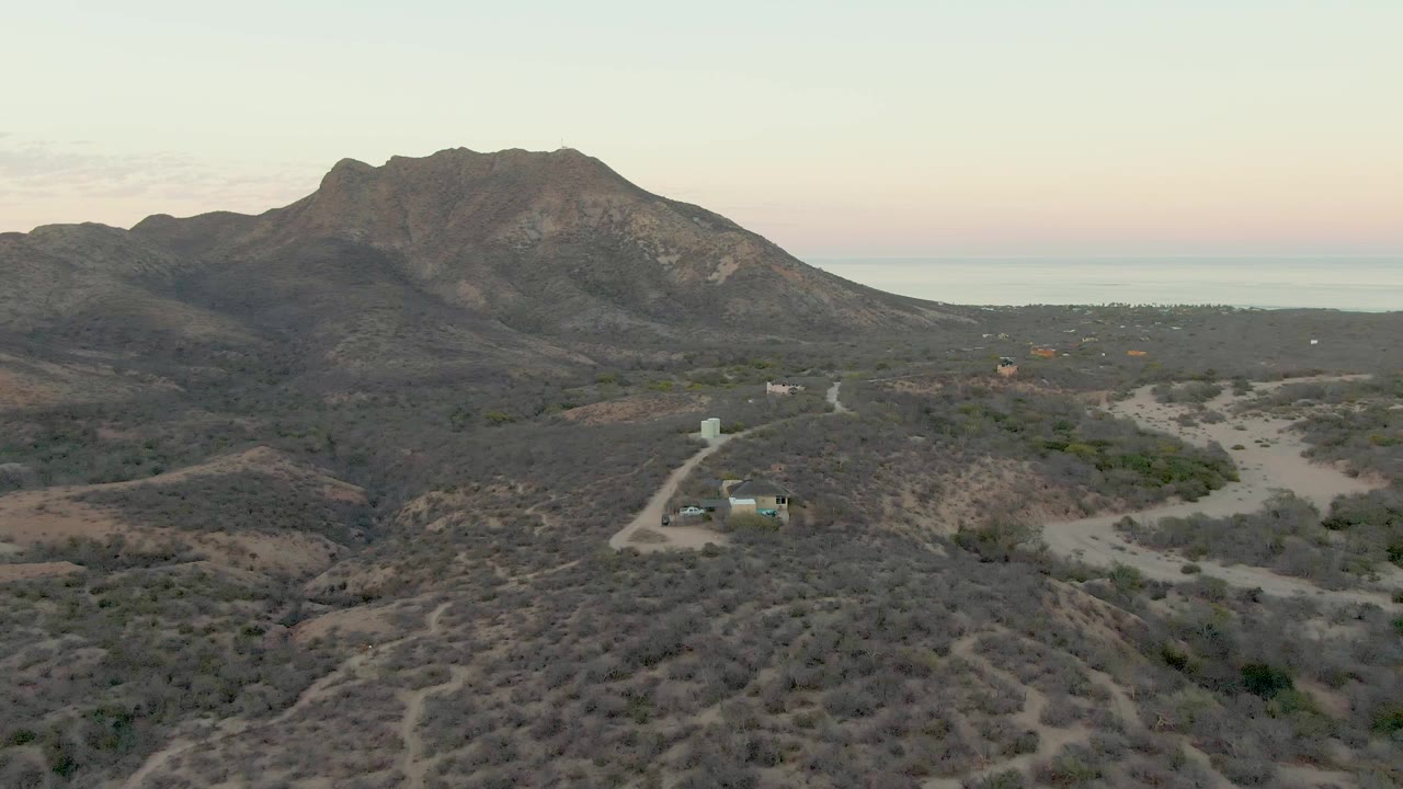 Early frame of Cabo Pulmo desert terrain in Baja California Sur, Mexico, with scrubby hills, a small structure beside a dirt road, a mountain ridge beyond, and no visible people.