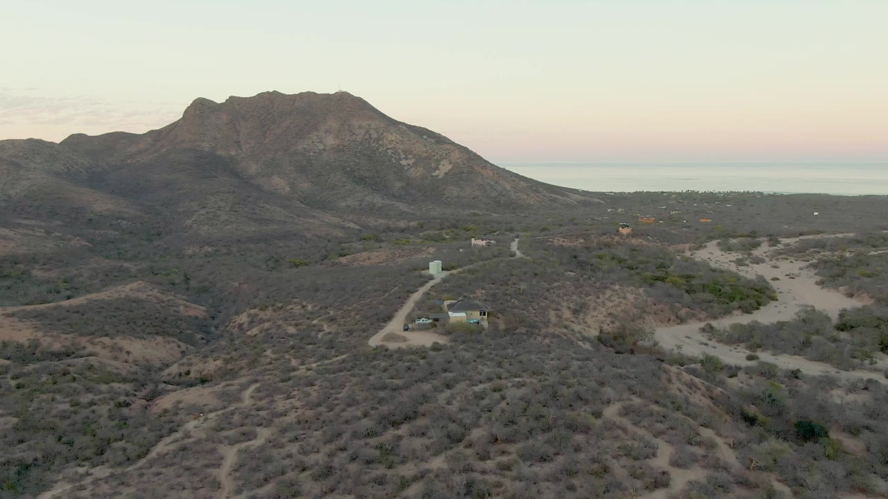 Cabo Pulmo aerial view one second into the clip, showing dry shrubland in the foreground, a small building and road in the midground, a broad mountain ridge, and no visible people.