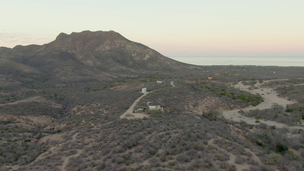 Mid-early frame from Cabo Pulmo, Baja California Sur, with winding dirt tracks through arid hills, a small structure left of center, the mountain ridge behind it, and no visible people.