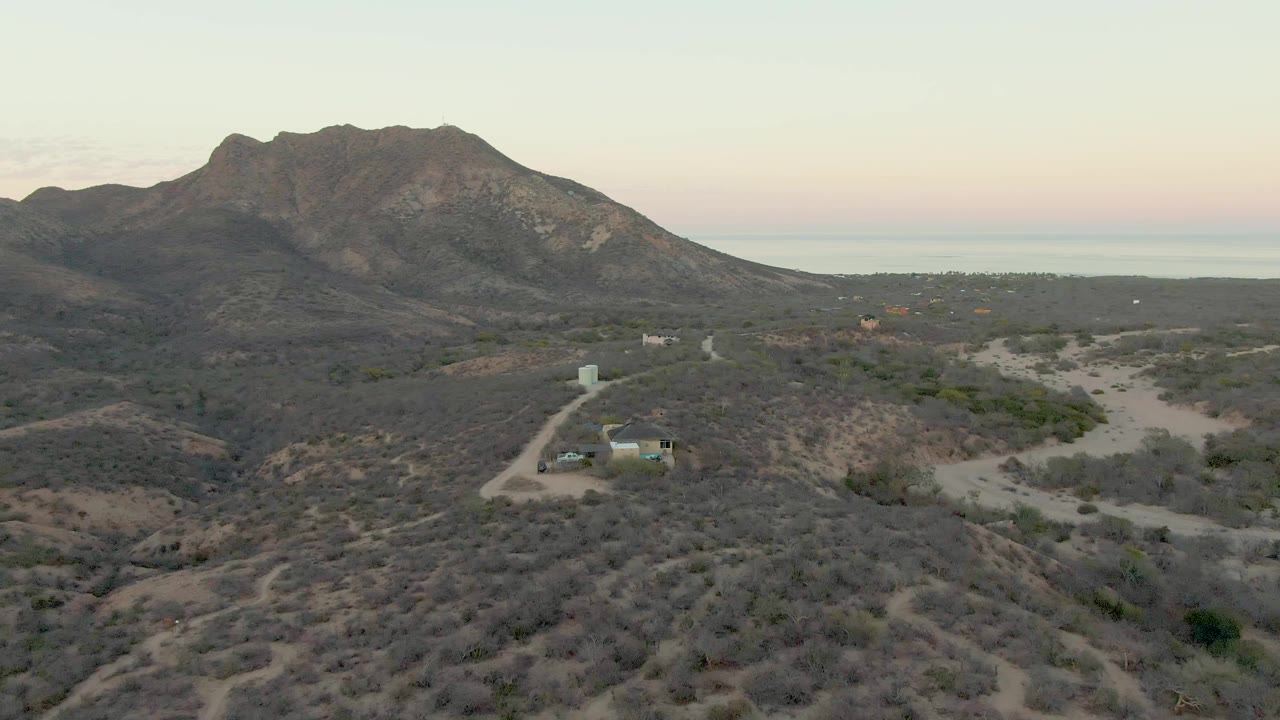 Desert slope with scrub, dirt road, small structure, and mountain under a pale sky.