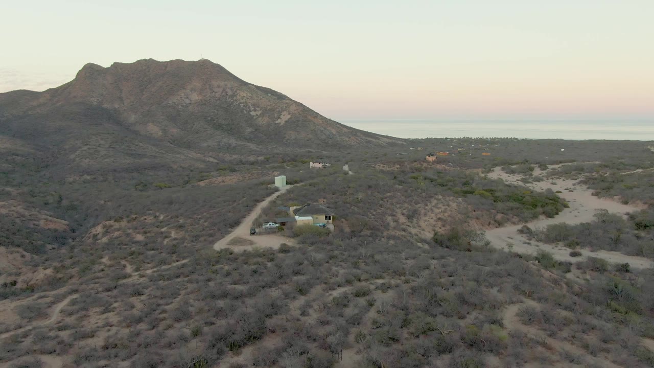 Wide aerial view in Cabo Pulmo, Baja California Sur, showing a car on a pale dirt road, dry scrubland, a small structure, a mountain, and no visible people.