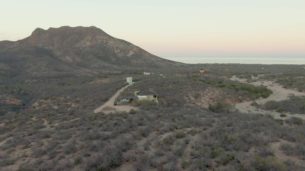 Elevated view over Cabo Pulmo scrubland showing the car on the dirt access road, a small building or structure, a broad mountain ridge, distant water or horizon line, and no visible people.