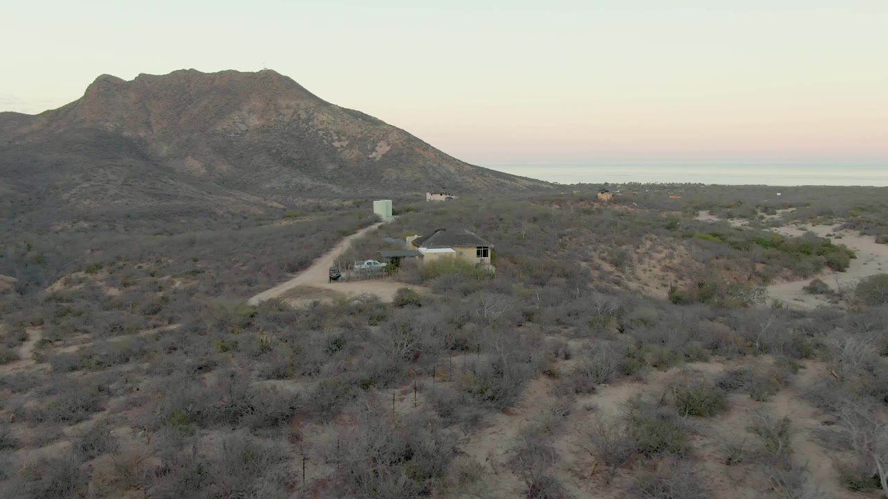 Opening elevated view in Cabo Pulmo shows a small desert house beside a dirt road with a parked vehicle, low scrub, a dark mountain ridge, pastel horizon light, and no visible people.