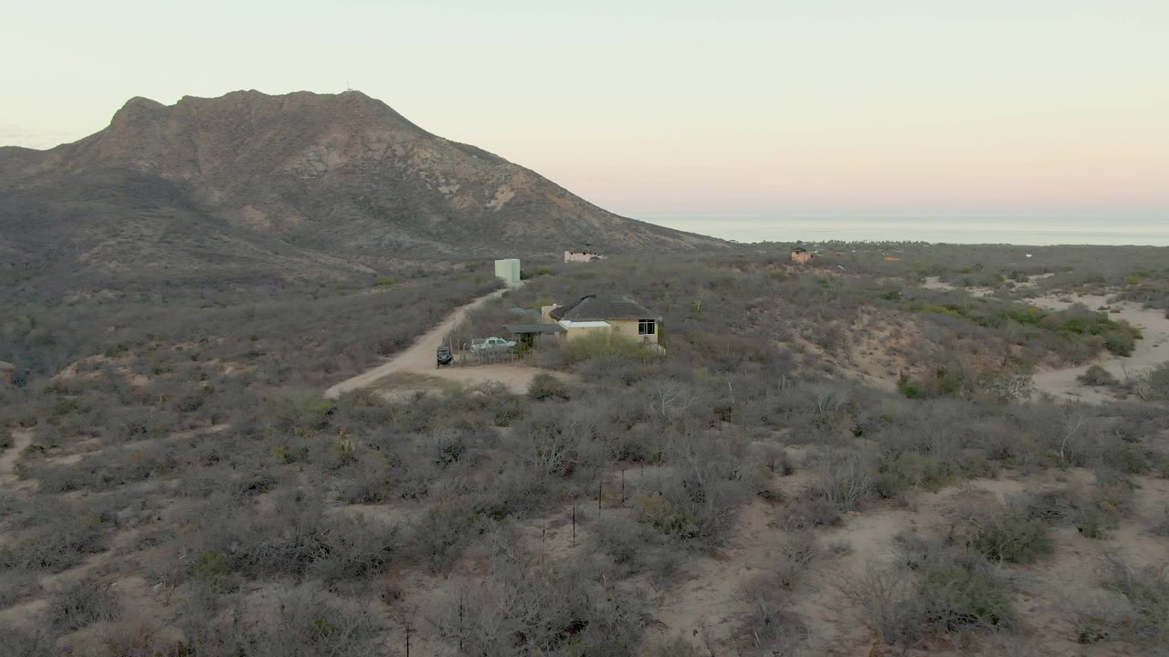 At 0:01, the Cabo Pulmo desert house remains in the midground beside the pale road, with scrub-covered foreground, the mountain ridge filling the upper frame, and no visible people.