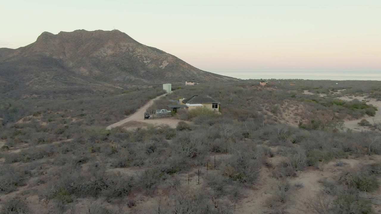 At 0:02, the elevated Cabo Pulmo view centers the house, vehicle, and dirt approach road against gray-green desert brush and a broad mountain backdrop, with no visible people.
