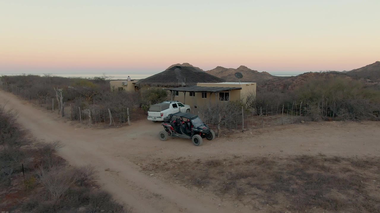 Wide aerial view in Cabo Pulmo, Baja California Sur with a white pickup near the left side of a dirt road, a small house, a parked ATV, dry brush, low hills, and no visible people.