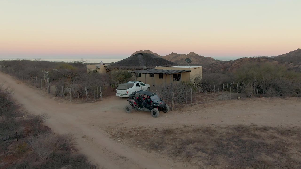 Aerial frame from Cabo Pulmo, Baja California Sur showing the white pickup closer to the small house, with the ATV parked in front, scrubby desert vegetation, distant hills, and no visible people.