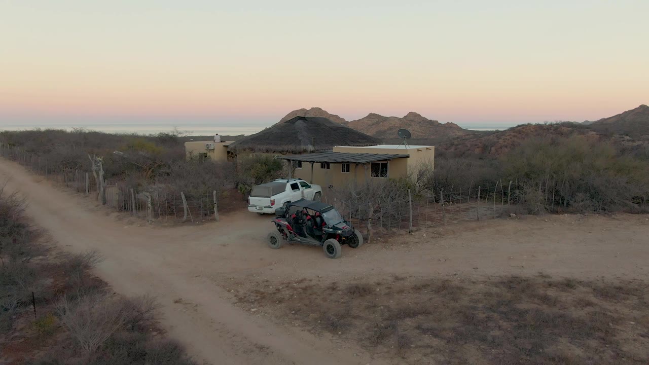 Wide drone-style view in Cabo Pulmo, Baja California Sur with the white pickup passing the house and parked ATV, a dirt road crossing the foreground, dry brush around the property, and no visible people.