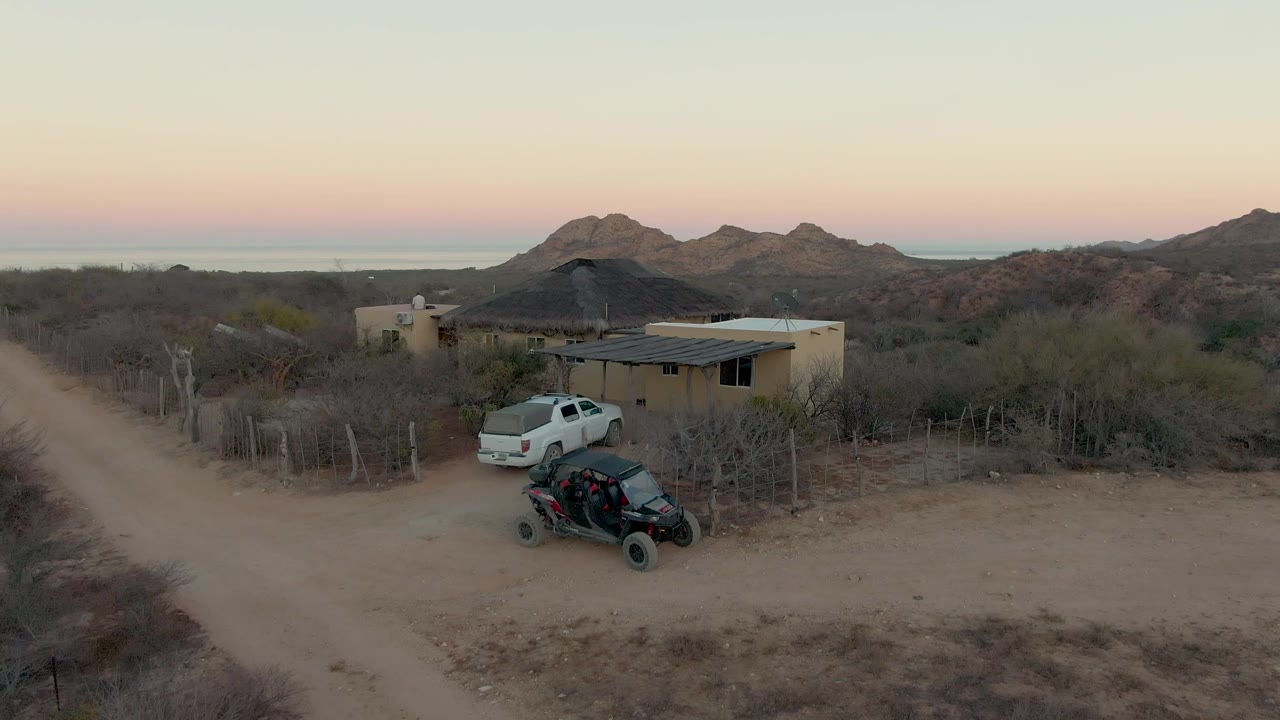 Aerial frame in Cabo Pulmo showing a tan desert house, white truck, black off road vehicle, dirt road, dry brush, distant mountains, pink orange sky, and no visible people.