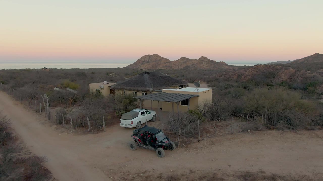 Aerial frame in Cabo Pulmo showing the house beside a winding dirt road, a white truck near the wall, a black off road vehicle in the foreground, dry vegetation, mountains, and no visible people.