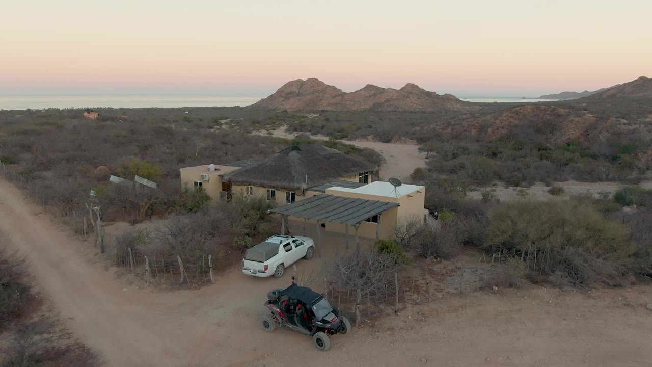 Aerial dusk frame in Cabo Pulmo, Baja California Sur, showing a tan desert house beside a dirt road, a white car near the carport, a black off-road vehicle in front, dry scrub, mountains, and no visible people.