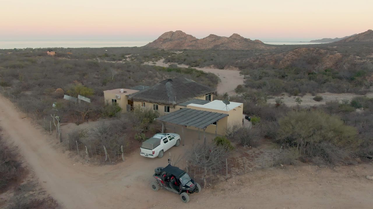 Cabo Pulmo aerial frame showing the tan house centered near a dirt track, a white car by the covered parking area, a black off-road vehicle in the foreground, dry vegetation, distant hills, and no visible people.