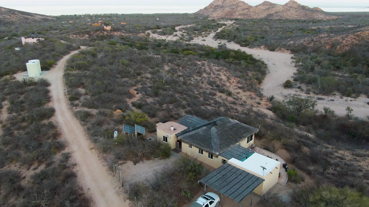 High aerial view in Cabo Pulmo, Baja California Sur showing a tan desert house, dirt road, pale water tank, scrub hills, distant mountains, and no visible people.