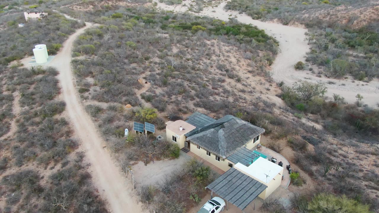 Drone footage over Cabo Pulmo, Baja California Sur frames the house lower right with a sandy road, water tank, dry brush, low hills, and no visible people.
