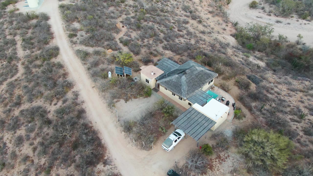 Aerial frame over a desert house in Cabo Pulmo, Baja California Sur, showing ground solar panels, a dirt road, a covered driveway, a white parked vehicle, sparse vegetation, and no visible people.