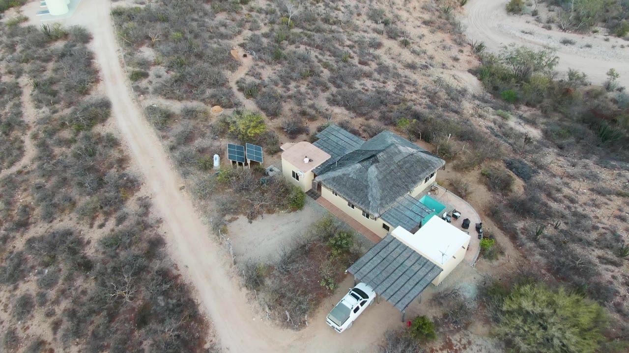 Drone view over the Cabo Pulmo desert property with the house slightly larger in frame, solar panels left of the roof, a white vehicle under the driveway cover, and no visible people.
