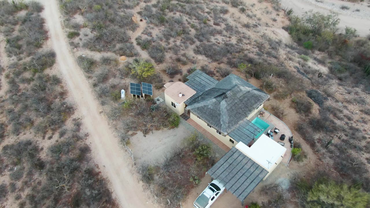Overhead frame in Cabo Pulmo, Baja California Sur, with the beige house, dark roof, solar panels, dirt road, covered driveway, white vehicle, and surrounding dry brush visible with no people in frame.