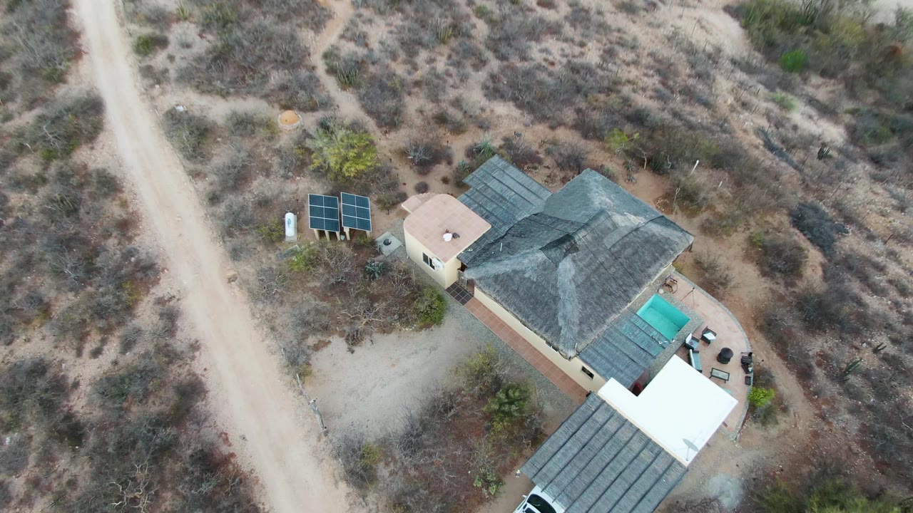 Closer aerial view of the desert house roof, solar panels, dirt road, and parked vehicle.