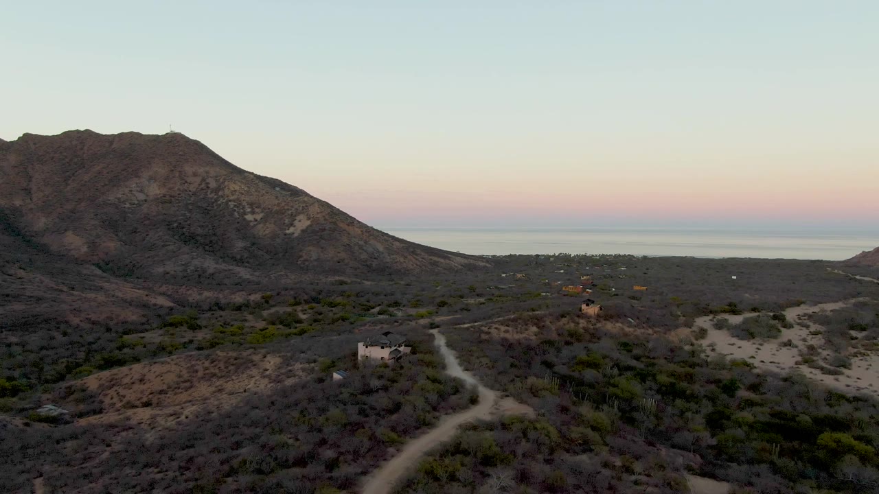 At 0:00, a wide drone view in Cabo Pulmo shows pale cylindrical tanks near a winding dirt road, scrubby desert hills, a large mountain, distant sea horizon, pastel dusk sky, and no visible people.
