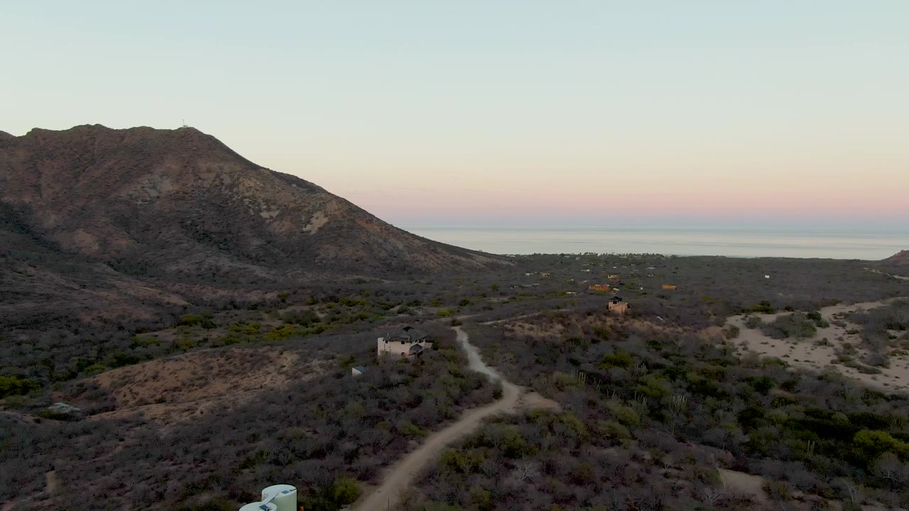 At 0:01, the Cabo Pulmo aerial view keeps the tanks low in frame while the dirt road curves through dry vegetation toward scattered buildings, the mountain, the sea horizon, and no visible people.