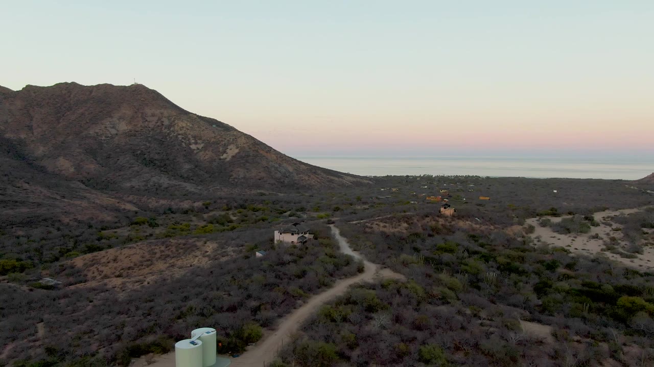 At 0:02, the drone composition shows the pale tanks near the bottom edge, the dirt road splitting across the desert floor, a few buildings, the mountain profile, the distant sea, and no visible people.