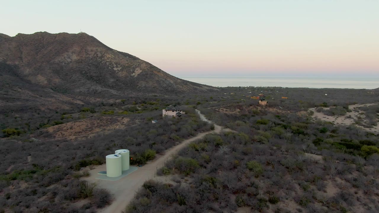 A high aerial view over Cabo Pulmo desert brush shows a dirt road, pale green water tanks in the lower left, distant small buildings, mountains, sea horizon, and no visible people.