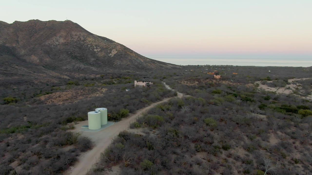 An aerial view in Cabo Pulmo shows the dirt track and water tanks slightly farther back in frame, with low desert vegetation, small buildings, mountains, a pale sea line, and no visible people.