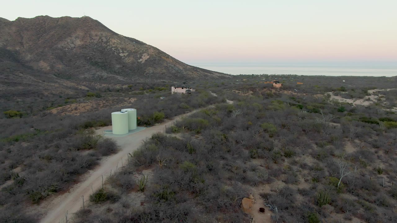 Aerial view in Cabo Pulmo, Baja California Sur, showing a dirt road beside two pale green water tanks, dry scrubland, scattered houses, mountains, and a soft sunset horizon.