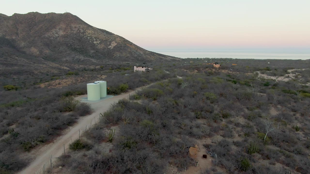 Drone view of a pale dirt road passing water tanks through dry desert brush toward mountains.