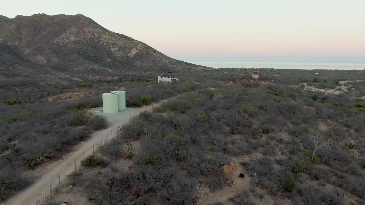 Wide drone view over Cabo Pulmo desert terrain, with the dirt road angling past two green tanks, scattered homes across the midground, mountains on the left, and a pale evening horizon.