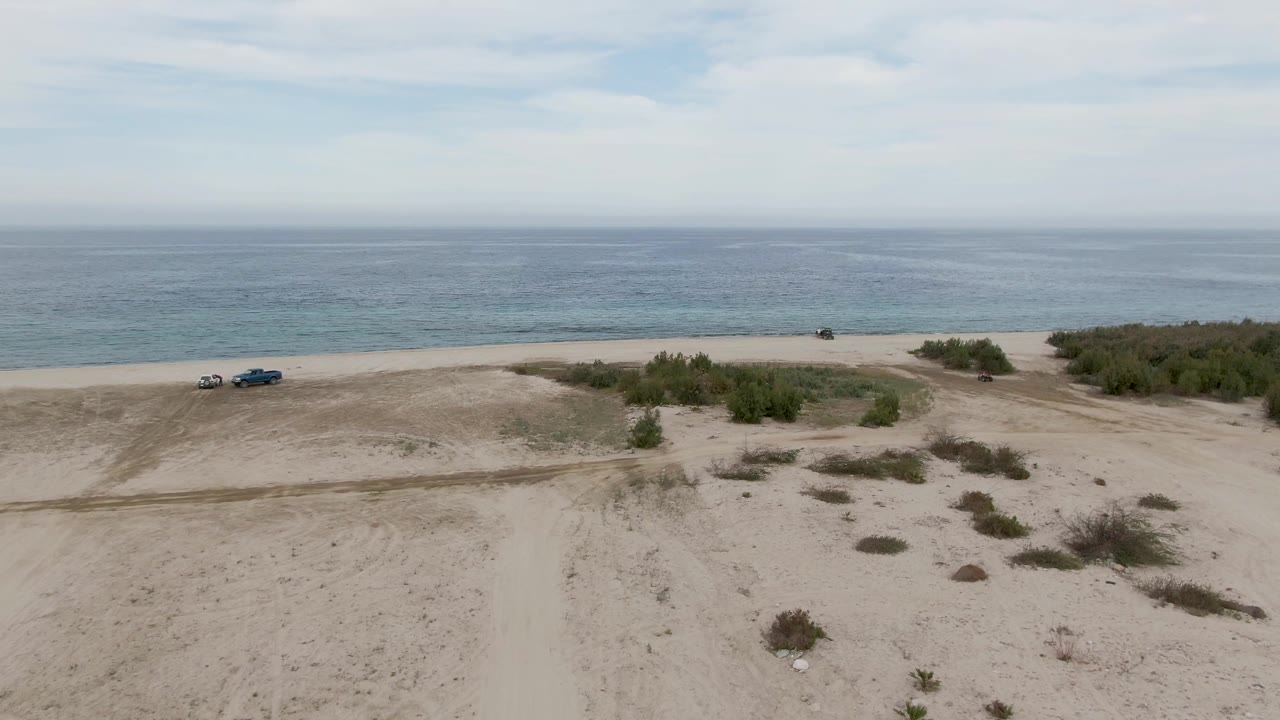 Aerial frame from Los Barriles, Baja California Sur, showing two vehicles near the left side of a sandy beach, low shrubs inland, blue ocean beyond, and no visible people.