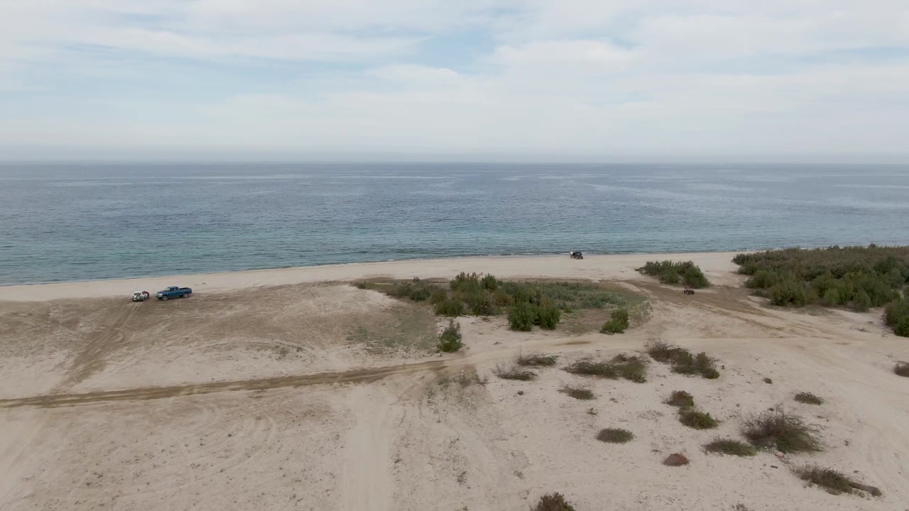 Vehicles drive along pale sand with ocean water behind them.