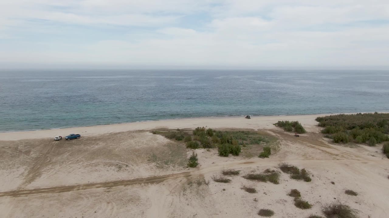 Aerial frame from Los Barriles, Baja California Sur, showing the vehicles near center-left on the sand, a larger shrub patch to the right, calm blue-green water in back, and no visible people.