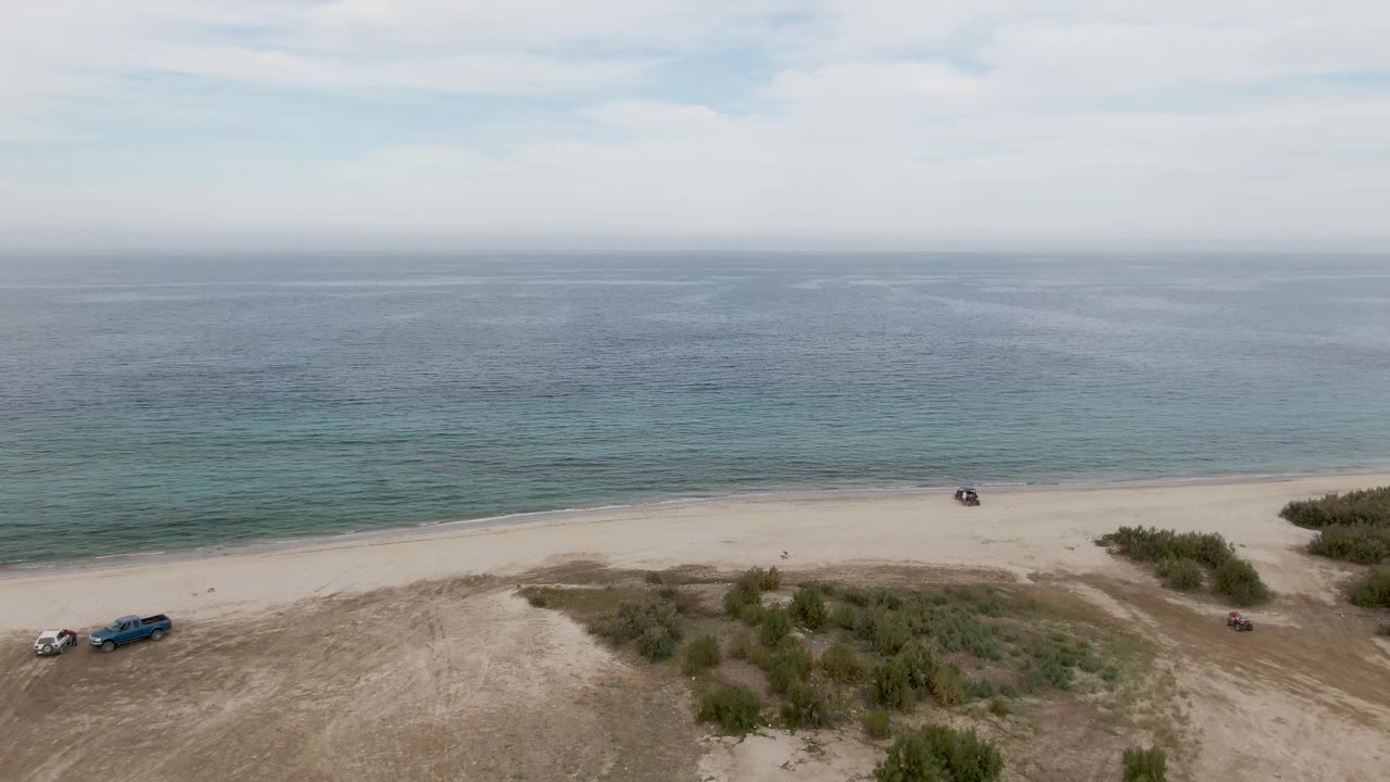 Aerial view of sandy beach, shrubs, vehicles, shoreline, and blue-green ocean