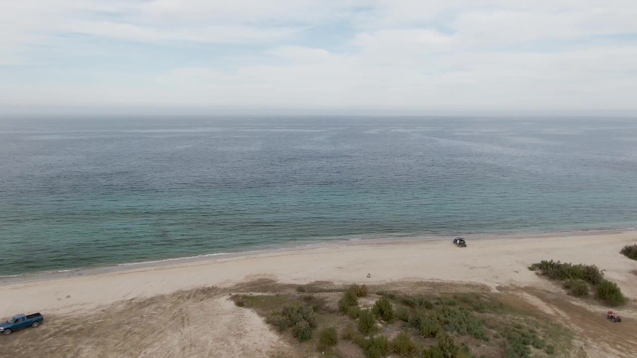 Aerial view in Los Barriles with a blue vehicle near the lower left, a smaller vehicle near beach shrubs, a dark vehicle by the waterline, pale sand, calm ocean, and no visible people.