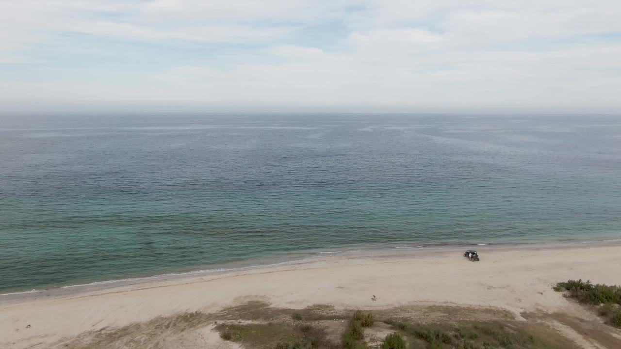 Wide aerial frame over Los Barriles showing a long band of pale sand, a dark vehicle near the waterline, shrubs at the right edge, blue-green ocean, overcast sky, and no visible people.