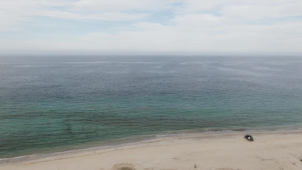 Los Barriles aerial frame with most of the view filled by textured blue-green ocean, a thin strip of sandy beach at the bottom, one dark vehicle near the shore, and no visible people.