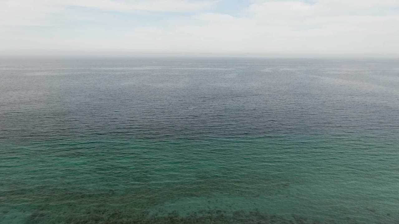 Aerial frame over Los Barriles water with the horizon still high in frame, blue-green ocean surface, light ripples, and darker seabed areas beginning to show through with no visible people.