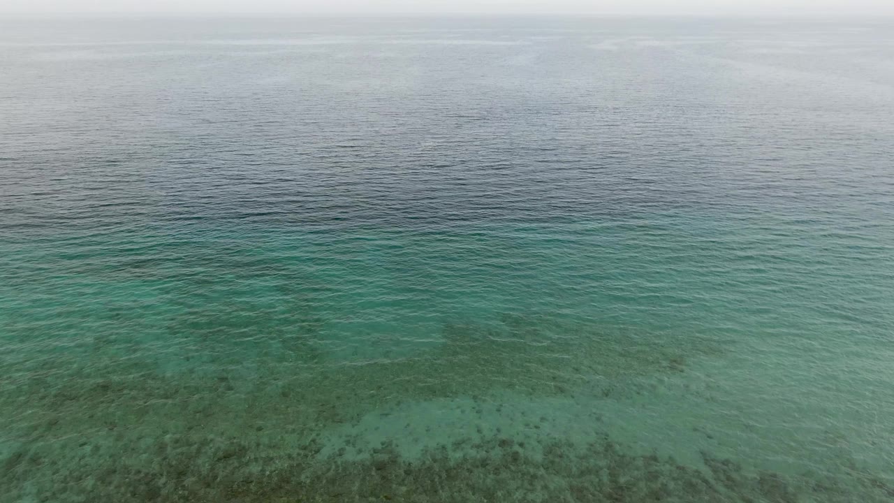 Aerial view above Los Barriles turquoise water where darker coral or seabed patches are visible beneath gentle ripples, with deep water and cloudy sky near the horizon and no visible people.