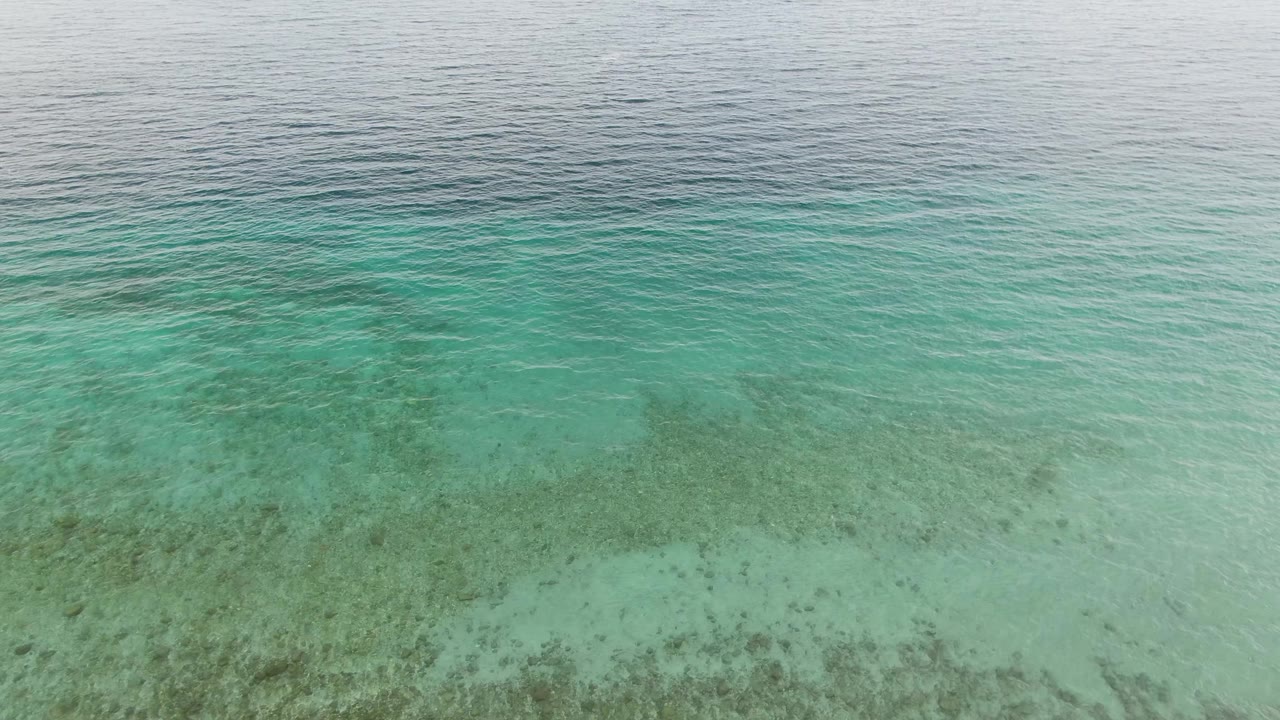 Low aerial frame over Los Barriles clear turquoise shallows, with pale sandy seabed across the lower frame, darker underwater patches through the middle, and no visible people.